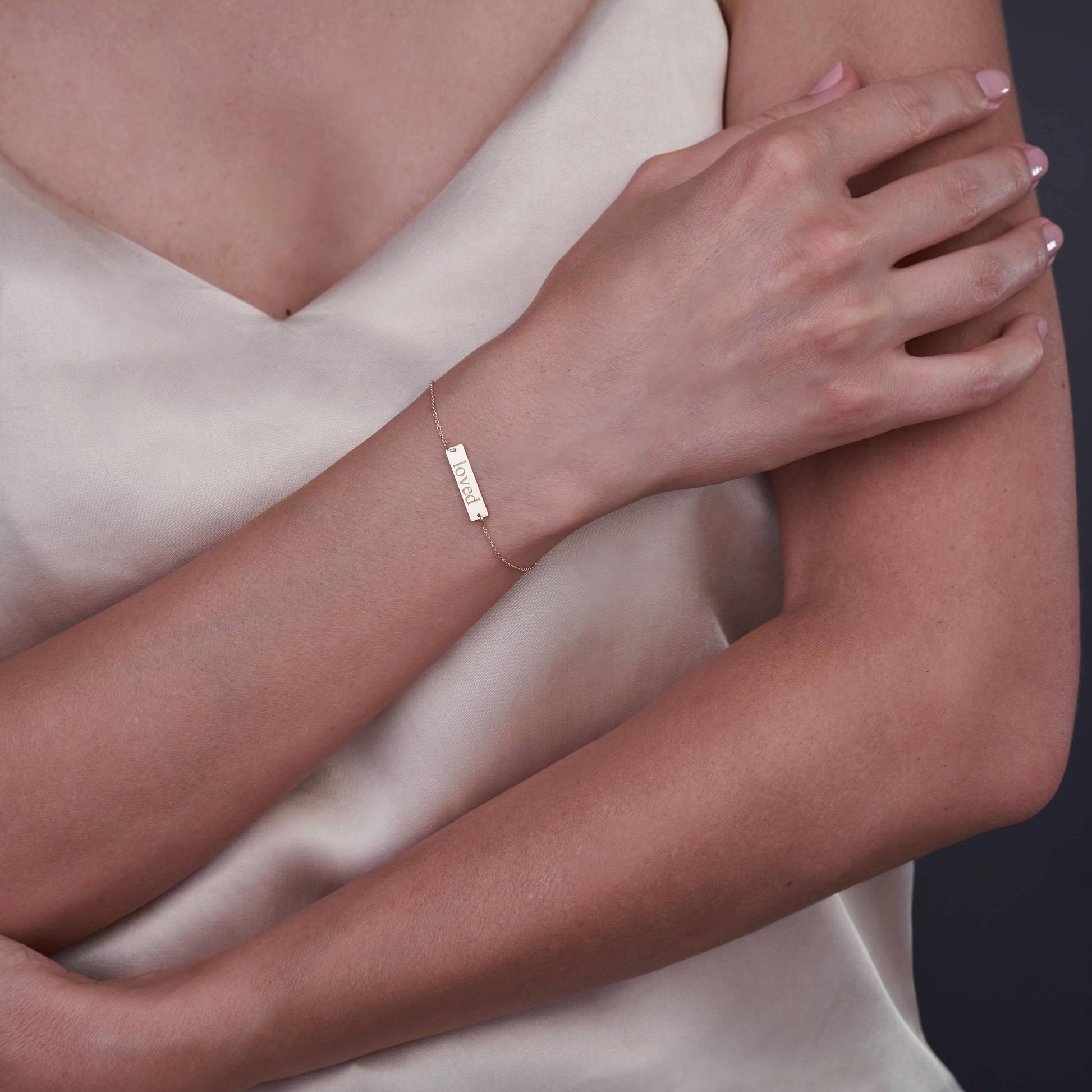 Близък план на човек, носещ златна гривна 14K с гравирана дума 'Loved', на неутрален фон.